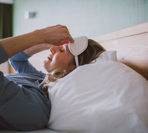 Das Bild zeigt eine Frau mit einem blauen langen Oberteil. Sie liegt in einem Bett, das weiß bezogen ist und zieht sich gerade eine weiße Schlafmaske an.