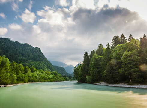 Das Bild zeigt eine friedliche Landschaft um den Fluss Lech, der durch einen grünen Wald fließt. Der Fluss, mit seinem leicht türkisenen Wasser, ist der zentrale Fokus des Bildes. Er windet sich durch den dichten Wald, erzeugt einen Sinn von Ruhe und Harmonie mit der Natur. Der Wald ist ein kräftiges Grün, gefüllt mit hohen Bäumen, die über den Fluss hineinwachsen. Der blaue Himmel darüber ist mit flauschigen weißen Wolken durchzogen, die zur Schönheit der Szene beitragen. Das Bild wird aus der Ferne aufgenommen, was einen umfassenden Blick auf den Fluss sowie den umliegenden Wald erlaubt. Die Perspektive des Bildes betont Tiefe, was die Weite des Waldes und des Flusses unterstreicht. Das Bild ist eine schöne Darstellung von Naturfrieden und der Harmonie zwischen dem Fluss und dem Wald.