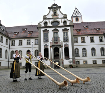 Die Szenerie zeigt den Innenhof des ehemaligen Benediktinerklosters St. Mang. Die weiße Fassade ist mit vielen Fenstern durchzogen. Der Mittelteil des Gebäudes ist höher und mit Illusionsmalerei verziert. Im Vordergrund stehen ein Mann und zwei Frauen, die Alphorn spielen. Die Personen sind in Tracht gekleidet. Der Boden ist mit Kopfsteinpflaster bedeckt. 