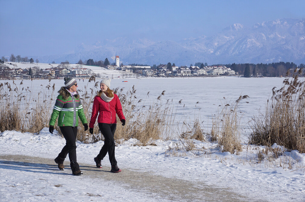 Das Bild fängt eine ruhige Winterlandschaft ein. Zwei Frauen sind auf einem schneebedeckten Uferweg zu sehen. Der Weg, auf dem sie gehen, ist von hohen Gräsern gesäumt. Der See ist zugefrohren. Im Hintergrund ist der Ort Hopfen zu erkennen. Jenseits der Ortschaft erheben sich majestätische Berge, deren Gipfel mit Schnee bedeckt sind. Der Himmel ist blau und sonnig.