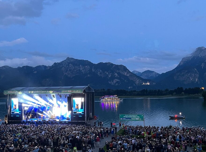 Das Foto zeigt eine große, hell erleuchtete Open Air Bühne am Ufer des Forggensees. Im Vordergrund stehen unzählige Personen. Im Hintergrund sind die Berge und das beleuchtete Schloss Neuschwanstein zu sehen, davor liegt der Forggensee auf dem ein beleuchtetes Schiff fährt.  