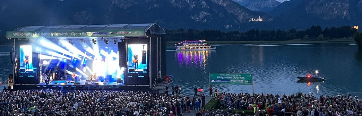 Das Foto zeigt eine große, hell erleuchtete Open Air Bühne am Ufer des Forggensees. Im Vordergrund stehen unzählige Personen. Im Hintergrund sind die Berge und das beleuchtete Schloss Neuschwanstein zu sehen, davor liegt der Forggensee auf dem ein beleuchtetes Schiff fährt.  