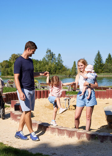 ie Szenene in dem Bild ist eine friedliche Momentaufnahme in einem Park. Vor einem ruhigen See stehen ein Mann und eine Frau, beide tragen legeres Outfit. Der Mann hat ein blaues T-Shirt und blaue Shorts an, während die Frau ein weißes T-Shirt und blaue Shorts trägt. Sie halten Händchen, was ihre Nähe zum Ausdruck bringt. Im Hintergrund ist ein friedlicher Teich zu sehen, das zur friedlichen Atmosphäre des Parks beiträgt. Einige Bäume stehen dort und bieten eine natürliche Kulisse für die Szene. Der Park ist auch mit einem Spielplatz ausgestattet, inklusive eines Sandkastens und eines Schaukelsatzes. Der Sandkasten liegt nahe zum See, bietet also eine fröhliche und aktive Spielmöglichkeit für Kinder. Der Schaukelsatz befindet sich etwas weiter weg, wo Kinder ungetehrt spielen und Eltern ihnen zuschauen können. Das Bild zeigt generell einen typischen Tag im Park, mit Leuten, die die Natur genießen, und Kindern, die im Sandkasten und auf den Schaukeln spielen.