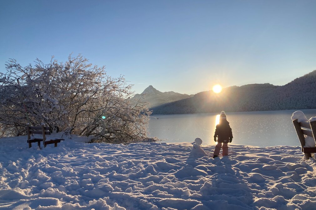 Das Bild zeigt eine beruhigende Winterlandschaft. Eine Person, gekleidet in einen schwarzen Mantel und eine weiße Mütze, steht auf einer mit Schnee bedeckten Fläche. Sie sind in der Mitte des Bildes positioniert und blicken in Richtung des rechten Randes. Die Person scheint auf die majestätischen Berge zu schauen, die in der Ferne aufragen. Der Himmel darüber ist ein klarer blauer Ton, der einen beeindruckenden Kontrast zum weißen Schnee bietet. Der Boden ist mit Schnee bedeckt, mit ein paar Bäumen verstreut herum. Die gesamte Szene vermittelt ein Gefühl von Ruhe und Frieden.