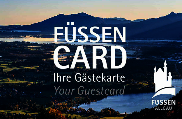 Das Bild zeigt eine Karte, in der Größe einer Scheckkarte. Diese ist mit einem Panoramafoto bedruckt und trägt die Aufschrift „Füssen Card, Ihre Gästekarte, Your guestcard“ Im Hintergrund sieht man unscharf bunte Häuserfassaden. Der Himmel ist blau. Die Sonne scheint.