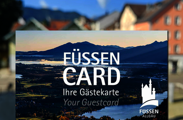 Das Bild zeigt eine Karte, in der Größe einer Scheckkarte. Diese ist mit einem Panoramafoto bedruckt und trägt die Aufschrift „Füssen Card, Ihre Gästekarte, Your guestcard“ Im Hintergrund sieht man unscharf bunte Häuserfassaden. Der Himmel ist blau. Die Sonne scheint.