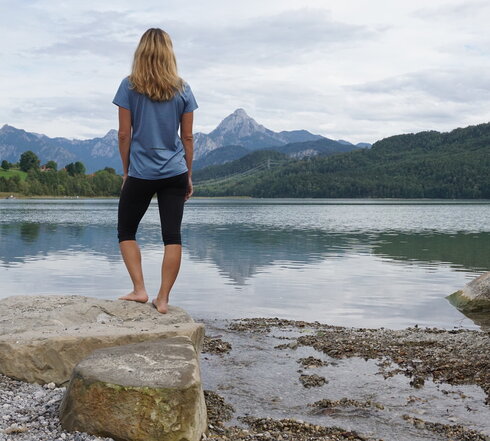 Ein See mit einem bewaldeten Ufer wird von einem Felsvorsprung und einer Person, die barfuß steht, dominiert. Im Hintergrund erheben sich Berge.