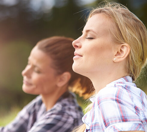 Das Bild zeigt zwei junge Frauen, die mit geschlossenen Augen das Gesicht in die Sonne halten und die Situation sichtlich genießen. Man sieht lediglich die Gesichter und einen Teil des Oberkörpers beider Frauen. Sie tragen karierete Oberteile und haben die Haare zusammengebunden. 