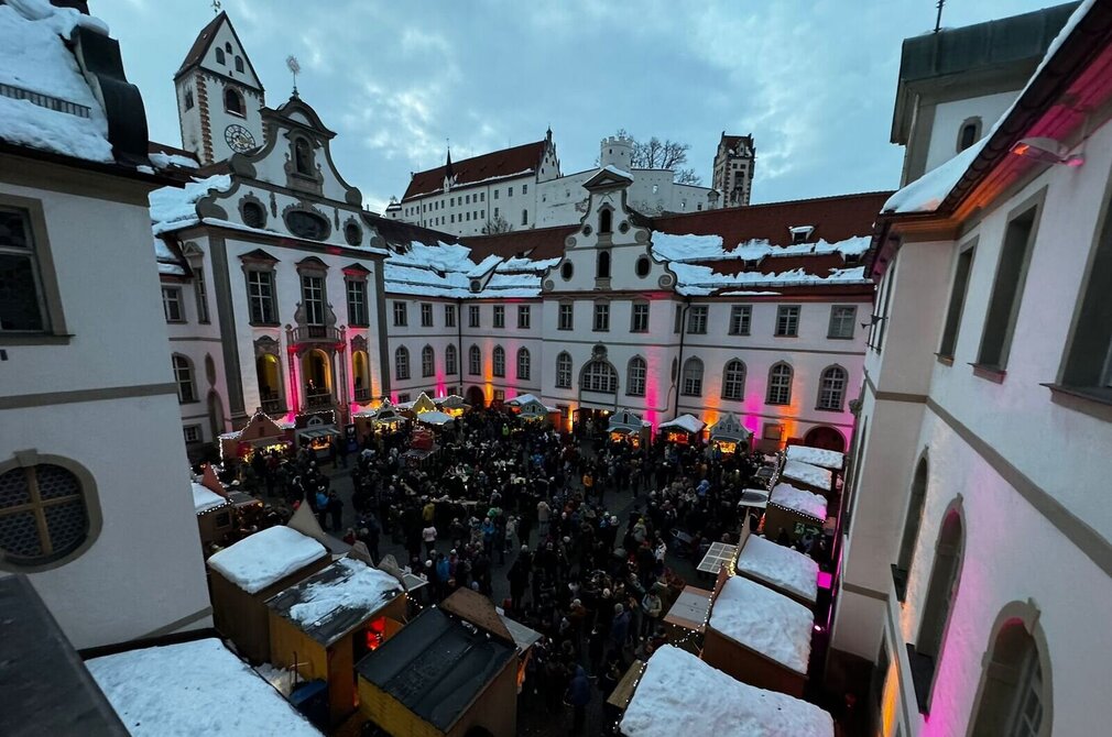 Das Bild zeigt den Füssener Adventsmarkt im Klosterhof des ehemaligen Benediktinerklosters St. Mang. Die Fassade des rundumlaufenden Gebäudes ist pink beleuchtet. Die Buden des Adventsmarkt sind ebenfalls festlich beleuchtet. Zahlreiche Menschen sind zu sehen. Es dämmert, ist aber noch hell. Der Himmel ist wolkenverhangen.   