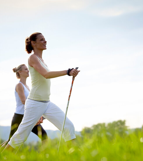 Das Bild zeigt zwei sportlich gekleidete, schlanke Frauen, die mit Nordic-Walking-Stöcken über eine grüne Wiese laufen. Beide Frauen haben die Haare zusammengebunden und blicken glücklich voraus. Der blaue Himmel ist von sanften Wolken durchzogen.