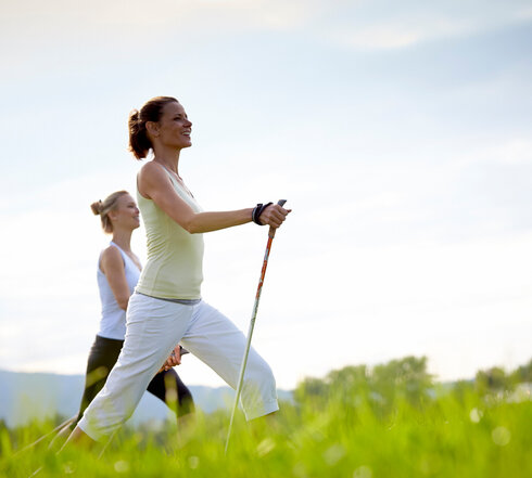 Das Bild zeigt zwei sportlich gekleidete, schlanke Frauen, die mit Nordic-Walking-Stöcken über eine grüne Wiese laufen. Beide Frauen haben die Haare zusammengebunden und blicken glücklich voraus. Der blaue Himmel ist von sanften Wolken durchzogen.