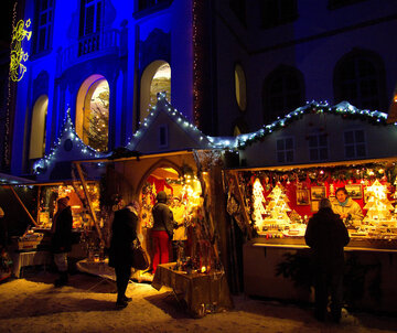 Das Bild zeigt beleuchtete Verkaufsbuden eines Weihnachtsmarktes. Menschen laufen umher und betrachten die Waren in den Buden. Die Verkäuferinnen beraten ihre Kunden. Man sieht viele Weihnachtsdekorationen in den Buden stehe. Im Hintergrund erhebt sich ein prachtvolles Gebäude, das blau beleuchtet ist. Es ist bereits dunkel. Der Boden ist schneebedeckt. 