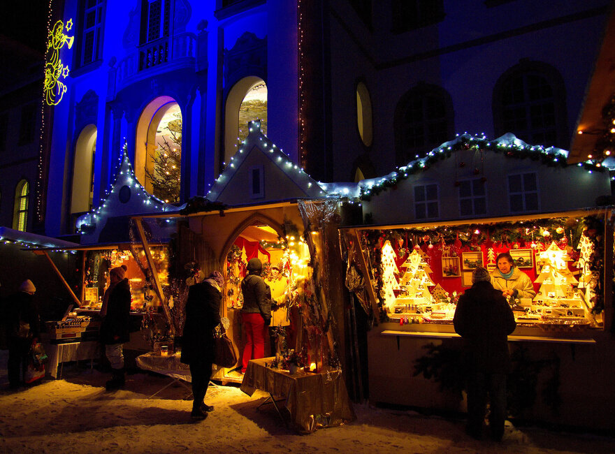 Das Bild zeigt beleuchtete Verkaufsbuden eines Weihnachtsmarktes. Menschen laufen umher und betrachten die Waren in den Buden. Die Verkäuferinnen beraten ihre Kunden. Man sieht viele Weihnachtsdekorationen in den Buden stehe. Im Hintergrund erhebt sich ein prachtvolles Gebäude, das blau beleuchtet ist. Es ist bereits dunkel. Der Boden ist schneebedeckt. 