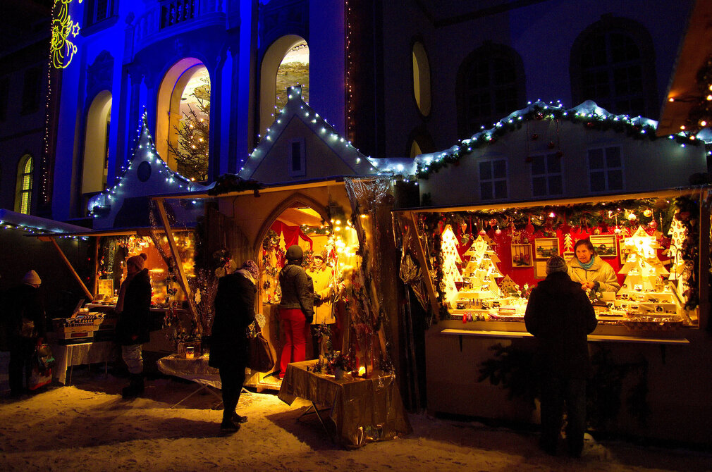 Das Bild zeigt beleuchtete Verkaufsbuden eines Weihnachtsmarktes. Menschen laufen umher und betrachten die Waren in den Buden. Die Verkäuferinnen beraten ihre Kunden. Man sieht viele Weihnachtsdekorationen in den Buden stehe. Im Hintergrund erhebt sich ein prachtvolles Gebäude, das blau beleuchtet ist. Es ist bereits dunkel. Der Boden ist schneebedeckt. 