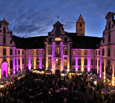 Das Bild zeigt den Füssener Adventsmarkt im Klosterhof des ehemaligen Benediktinerklosters St. Mang. Die Fassade des rundumlaufenden Gebäudes ist pink beleuchtet. Die Buden des Adventsmarkt sind ebenfalls festlich beleuchtet. Zahlreiche Menschen sind zu sehen. Gerade ist die Sonne untergegangen. Man sieht noch die letzten Sonnenstrahlen über dem Klosterdach.