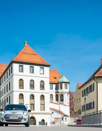 Ein silberner Mini fährt auf den Betrachter zu. Er fährt über eine Brücke. Im Hintergrund erhebt sich neben den bunten Altstadthäusern, der Gebäudekomplex des ehemaligen Benediktinerklosters mit seinen roten Dächern und unzähligen Fenstern. Der Himmel ist blau. Die Sonne scheint.