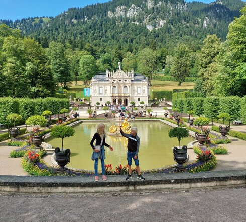 Das Bild fängt eine idyllische Szene in einem Park ein. Im Vordergrund stehen zwei Personen auf einer Steinmauer und blicken auf einen großen Teich. Der Teich ist von üppiger Vegetation umgeben, mit Bäumen und Sträuchern, die die Landschaft prägen. Ein Springbrunnen mit einer Statue befindet sich in der Mitte des Teichs und verleiht der Szene eine elegante Note. Im Hintergrund ragt ein majestätisches Burg-ähnliches Gebäude hoch, dessen Großzügigkeit durch die umgebende Vegetation betont wird. Der Himmel darüber ist ein klarer Blau, der auf einen angenehmen Tag hindeutet. Die Gesamtatmosphäre des Bildes ist eine der Ruhe und Schönheit.