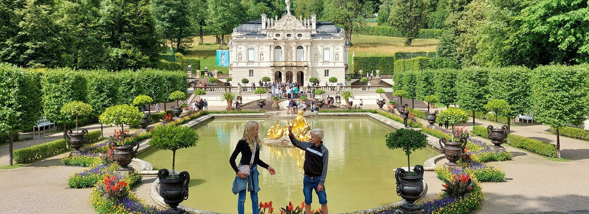 Das Bild fängt eine idyllische Szene in einem Park ein. Im Vordergrund stehen zwei Personen auf einer Steinmauer und blicken auf einen großen Teich. Der Teich ist von üppiger Vegetation umgeben, mit Bäumen und Sträuchern, die die Landschaft prägen. Ein Springbrunnen mit einer Statue befindet sich in der Mitte des Teichs und verleiht der Szene eine elegante Note. Im Hintergrund ragt ein majestätisches Burg-ähnliches Gebäude hoch, dessen Großzügigkeit durch die umgebende Vegetation betont wird. Der Himmel darüber ist ein klarer Blau, der auf einen angenehmen Tag hindeutet. Die Gesamtatmosphäre des Bildes ist eine der Ruhe und Schönheit.