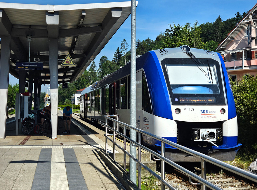 Das Bild zeigt einen Zug, der an einem Bahnsteig steht. Man sieht das Führerhaus des Zugs. Der Zug ist weiß-blau. Der Bahnsteig ist überdacht. Der Himmel ist blau. Die Sonne scheint.