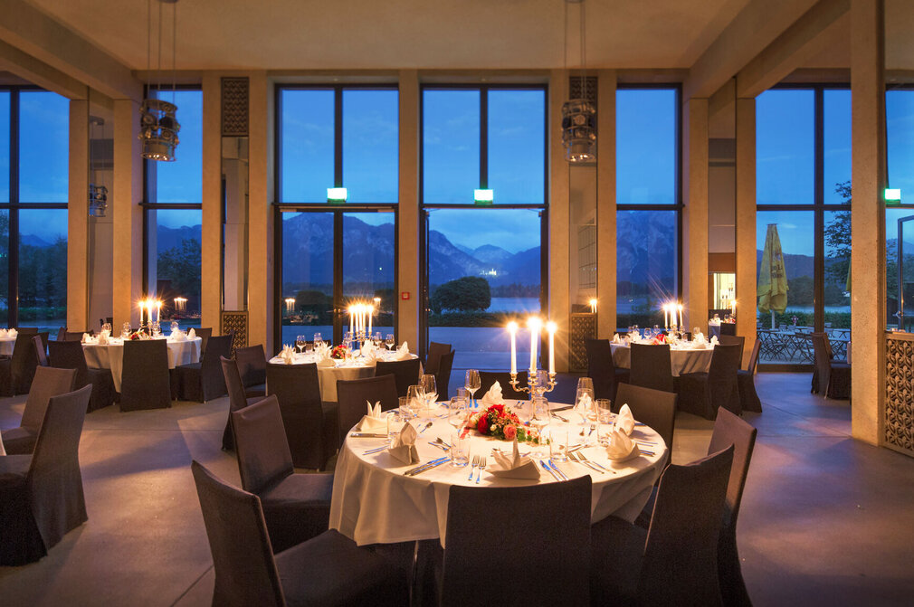 Das Foto zeigt einen großen Saal, in dem viele runde Tische stehen. Auf jedem Tisch liegt eine weiße Tischdecke. An jedem Tisch stehen 8 Stühle, die mit dunkelgrauen Hussen bezogen sind. Die Tische sind mit Gläser, Besteck, Blumenschmuck und Kerzenständern gedeckt. Durch die Fensterfront des Raumes hat man einen direkten Blick auf den Forggensee, die Berge und das Schloss Neuschwanstein. Es dämmert draußen. 