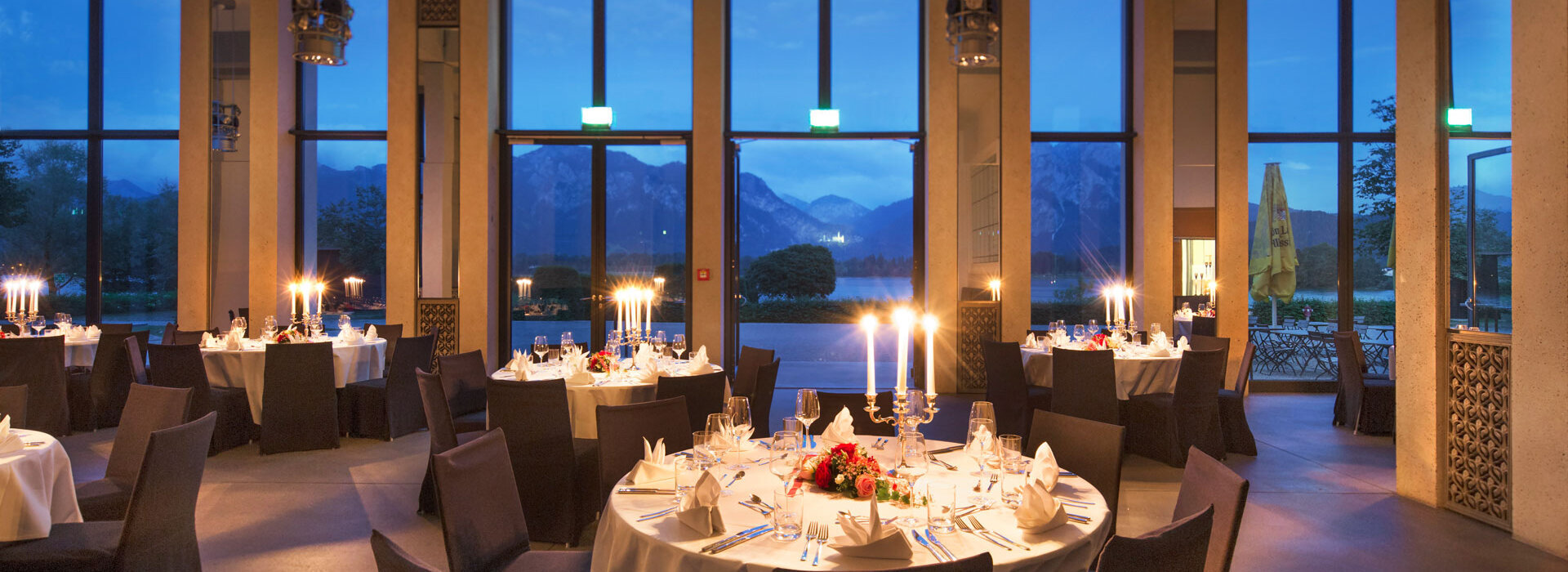 Das Foto zeigt einen großen Saal, in dem viele runde Tische stehen. Auf jedem Tisch liegt eine weiße Tischdecke. An jedem Tisch stehen 8 Stühle, die mit dunkelgrauen Hussen bezogen sind. Die Tische sind mit Gläser, Besteck, Blumenschmuck und Kerzenständern gedeckt. Durch die Fensterfront des Raumes hat man einen direkten Blick auf den Forggensee, die Berge und das Schloss Neuschwanstein. Es dämmert draußen. 