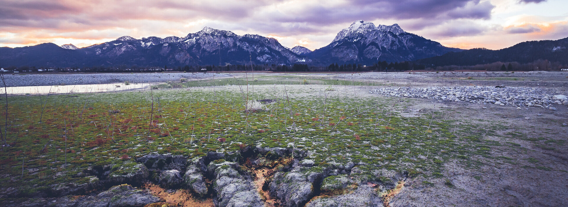 Forrgensee im Winter Das Bild fängt eine atemberaubende Aussicht eines gebirgigen Landschafts während des Sonnenuntergangs ein. Der Himmel ist mit verschiedenen Farben wie Orange, Pink und Purple angemalt und bildet eine beeindruckende Kulisse für die Szene. Die Berge, die im Schnee gehüllt sind, erheben sich majestätisch in der Ferne, ihre Spitzen berühren den Himmel