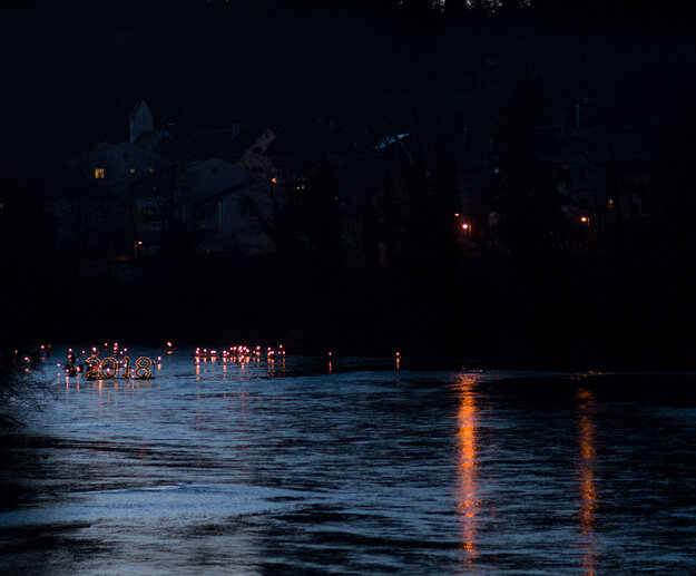 Das Bild zeigt einen dunklen Fluss. Über ihm sind die Umrisse von Häusern in der Dunkelheit zu erkennen. Auf dem Fluss schwimmt eine sehr große beleuchtete „2018“ im Hintergrund schwimmen zahlreiche Menschen mit brennenden Fackeln.   