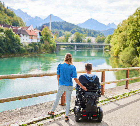 Das Bild fängt einen friedlichen Moment zweier Personen an einer Holzbrücke ein, die einen Blick auf eine Flusslandschaft bietet. Der Mann sitzt im Rollstuhl, während die Frau neben ihm steht. Im Hintergrund schmiegt sich ein malerisches Dorf in die üppige Vegetation.