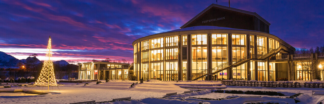 Das Foto zeigt das hellerleuchtete und verschneite Festspielhaus Neuschwanstein in der Abenddämmerung. Vor dem Festspielhaus steht eine Lichterkettenskulptur in Form eines Tannenbaums. Der Boden ist mit Schnee bedeckt. Der Himmel ist wolkenverhangen, die durch die Dämmerung teilweise eine rote Farbe angenommen haben.