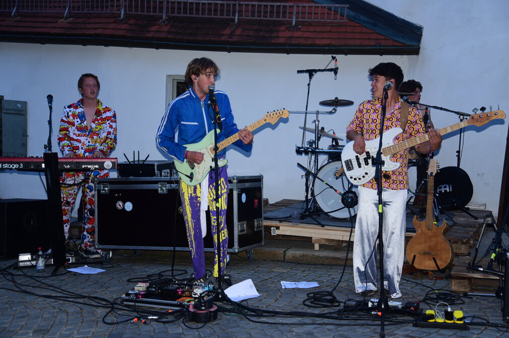 Das Bild zeigte vier Musiker, die bunte Kleidung tragen. Eine junger Mann spielt Keyboard, einer E-Gitarre, einer E-Bas und einer Schlagzeug. Diverse Kabel und Steckdosen sowie ein Mischpult liegen auf dem Kopfsteinpflaster vor ihnen. Im Hintergrund ist die weiße Fassade eines Hauses zu erkennen.  