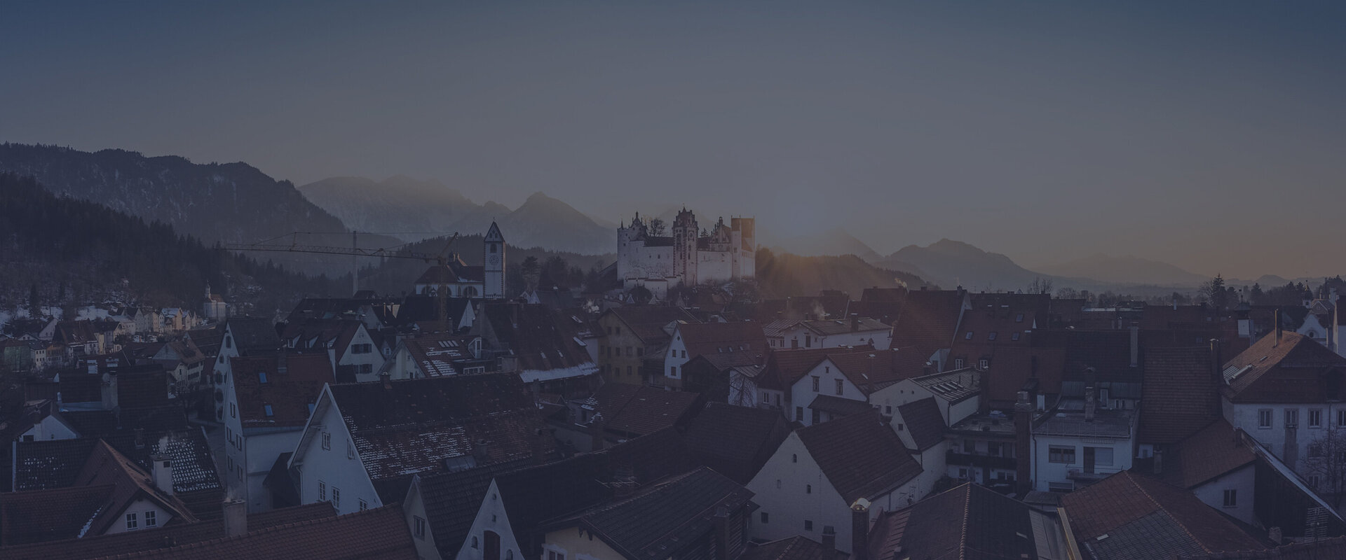 Hintergrundbild: Blick über die Dächer Füssens mit dem hohen Schloss und dem Kloster St. Mang im Sonnenuntergang