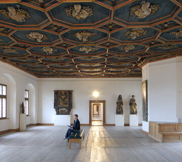 Der Raum hat eine hohe Decke mit einem dunkelblauen Deckenfresko und Holzdielen. Eine Person sitzt auf einer Bank, während Statuen und ein Gemälde im Raum ausgestellt sind.