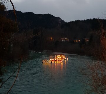 Das Bild zeigt einen dunklen Fluss. Im Hintergrund stehen Häuser am bewaldeten Ufer. Auf dem Fluss schwimmt eine sehr große beleuchtete „2024“ im Hintergrund schwimmen zahlreiche Menschen mit brennenden Fackeln.  