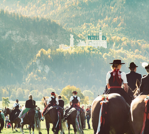 Das Foto zeigt ein große Anzahl von Reitern und Reiterinnen auf Pferden. Die Reiter tragen Tracht. Die Pferde sind festlich geschmückt. Im Hintergrund erhebt sich ein Berg, der mit einem grünen Bergwald überzogen ist. Auf einer Anhöhe vor dem Berg thront das weiße Schloss Neuschwanstein. 