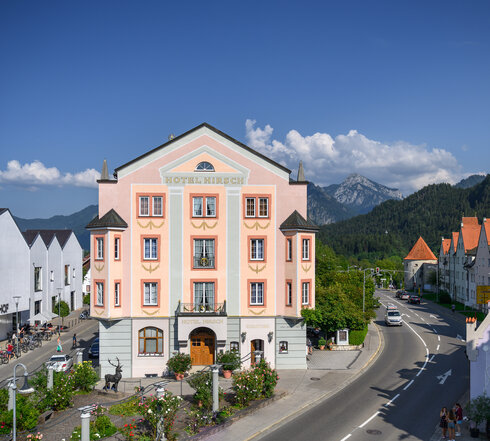 Das Bild zeigt eine beschauliche Stadt, die sich in die Berge schmiegt. Der Schwerpunkt des Bildes ist ein großes pinkfarbenes Gebäude, das sich vor dem Hintergrund der Berge abhebt. Das Gebäude ist mit einem Schild verziert, das "Hotel Mirch" lesen lässt, und signalisiert damit seine Funktion als Hotel. Die Straße vor dem Hotel ist von Bäumen gesäumt, die eine natürliche Note in die städtische Szene bringen. Der Himmel über dem Hotel ist ein klares Blau, was auf einen angenehmen Tag hindeutet. Die Berge im Hintergrund sind mit Schnee bedeckt, was der Szene einen winterlichen Touch verleiht. Das Gesamtbild zeichnet ein Bild einer friedlichen und idyllischen Stadt, die Besucher dazu einlädt, ihre Schönheit zu erkunden.