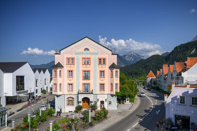 Das Bild zeigt eine beschauliche Stadt, die sich in die Berge schmiegt. Der Schwerpunkt des Bildes ist ein großes pinkfarbenes Gebäude, das sich vor dem Hintergrund der Berge abhebt. Das Gebäude ist mit einem Schild verziert, das "Hotel Mirch" lesen lässt, und signalisiert damit seine Funktion als Hotel. Die Straße vor dem Hotel ist von Bäumen gesäumt, die eine natürliche Note in die städtische Szene bringen. Der Himmel über dem Hotel ist ein klares Blau, was auf einen angenehmen Tag hindeutet. Die Berge im Hintergrund sind mit Schnee bedeckt, was der Szene einen winterlichen Touch verleiht. Das Gesamtbild zeichnet ein Bild einer friedlichen und idyllischen Stadt, die Besucher dazu einlädt, ihre Schönheit zu erkunden.