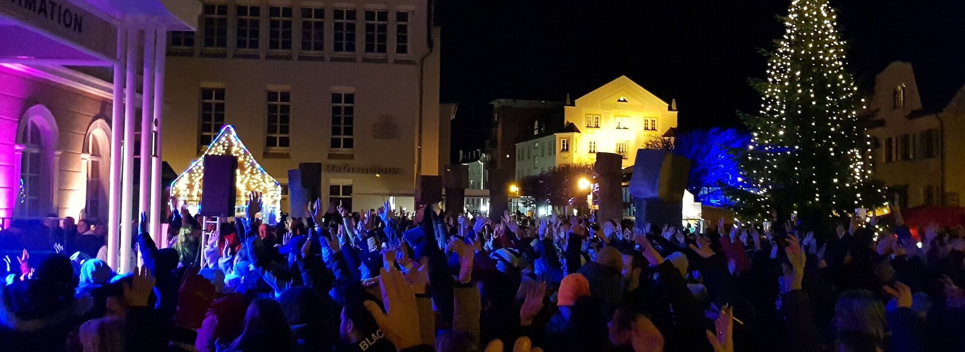 Das Bild zeigte einen Platz, um den viele Häuser stehen. Auf dem Platz stehen zahlreiche Menschen und strecken ihre Hände in de Höhe. Am einen Ende des Platzes steht ein beleuchteter Christbaum. Es ist dunkel.  