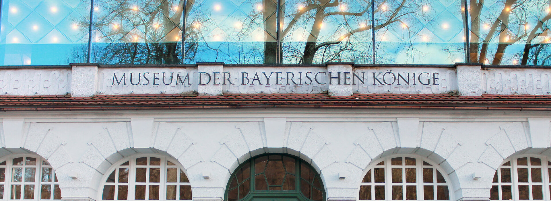 Das Bild zeigt einen großen, weißen Gebäudekomplex mit einer herausragenden Glasfassade. Das Gebäude ist mit einer Reihe von Bögen und Säulen verziert, was ihm einen Hauch von architektonischer Eleganz verleiht. Die Fassade wird von mehreren Fenstern unterbrochen, die von weißen Rolläden gerahmt sind. 