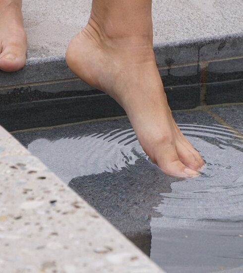 Das Bild eine Person, die vor einem Tretbecken steht und den linken Fuß ins Wasser streckt. Allerdings sind nur die nackten Füße der Person zu sehen.