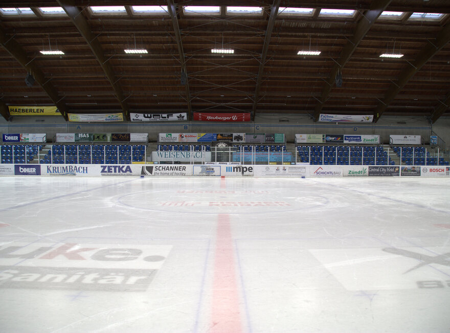 Man blickt von der Eisfläche eines Eisstadions auf die leere Zuschauertribüne. Die Decke des Stadions ist mit Lampen hell erleuchtet. An der Bande, den Wänden und unter der Eisfläche sind Werbeschilder zu sehen. Das Stadion ist menschenleer.