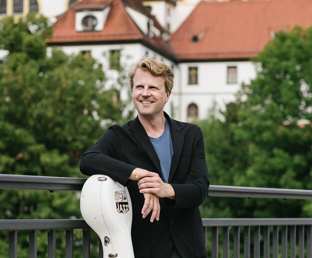 Auf dem Foto ist der Musiker Julian Steckel zu sehen, wie er mit einem Instrument auf der Theresienbrücke in Füssen steht.