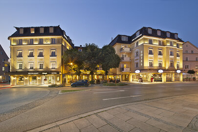  Das Bild fängt eine lebendige Stadtstraße in der Nacht ein, mit zwei großen Gebäuden, die prominent in der Ecke stehen. Das Gebäude auf der linken Seite ist ein Hotel, erkennbar an der großgeschriebenen Wort "HOTEL" auf der Fassade. Das Gebäude auf der rechten Seite ist ein Restaurant, wie durch das Wort "RESTAURANT" in großen Buchstaben angezeigt. Beide Gebäude sind beleuchtet, werfen ein warmes Licht auf die Straße unterhalb. Die Straße selbst ist mit Bäumen gesäumt, fügt der städtischen Kulisse eine Prise Natur hinzu. Ein paar Autos sind am Straßenrand geparkt und ein paar Fußgänger können auf dem Gehweg gesehen werden. Der Himmel über ist ein tiefes Blau, was darauf hinweist, dass es Nacht ist. Die Gesamtstimmung des Bildes ist lebhaft und einladend, fängt das Wesen des Stadtlebens in der Nacht ein.