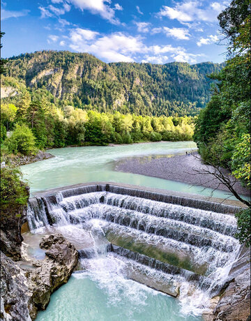 Das Bild zeigt einen atemberaubenden Blick auf einen Wasserfall – den Lechfall bei Füssen, der sich über eine felsige Klippe hinunterstürzt. Der Wasserfall, mit seinem weißen Schaum, ist der Hauptakzent im Bild. Er ist von üppig grünen Bäumen und Büschen umgeben, die der Szene eine Prise der Naturpracht verleihen. Der Wasserfall liegt in einem Tal, durch das der Fluss Lech fließt. Der klare blaue Himmel über ihm ist von fluffigen, weißen Wolken durchzogen. Die Perspektive des Bildes ist aus einer hohen Position, was eine Panoramaansicht des Wasserfalls und seiner Umgebung bietet.