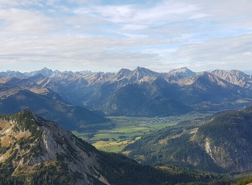 Man blickt auf unzählige Berggipfel, die sich vor dem Betrachter erheben. Dazwischen ist ein grünes Tal zu sehen. Dort liegt eine kleine Ortschaft. Der Himmel ist blau, jedoch von weißen Wolken durchzogen.