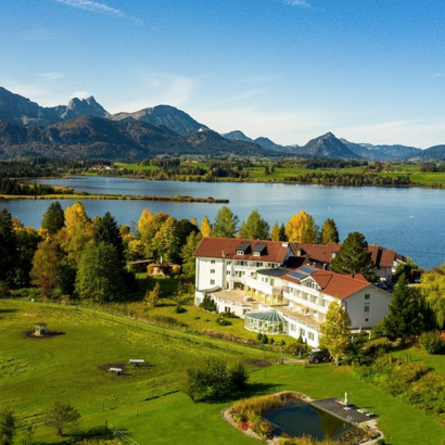 Das Bild zeigt ein großes weißes Haus, das umsäumt von Bäumen am Ufer eines Sees steht. Im Vordergrund sind grüne Felder und im Hintergrund die Gipfel der Allgäuer Alpen zu sehen.