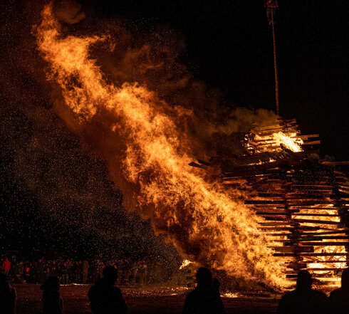 Das Foto zeigt ein großes brennendes Funkenfeuer. Das lichterloh mit einer großen Flamme brennt. Um das Feuer stehen viele Menschen und betrachten das Feuer. Es ist dunkel.  