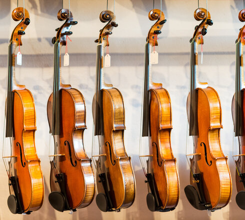 Lauten- und Geigenbau Das Bild zeigt eine Sammlung von Geigen, die jeder einzeln von einem Haken an einer Wand hängt. Die Geigen sind in einer Reihe angeordnet, mit jedem Instrument gleich in eine Richtung zeigend. Die Geigen sind unterschiedlicher Größe, was darauf hindeutet, dass sie für verschiedene Altersgruppen oder Spielweisen konzipiert sind. Die Geigen sind aus Holz und haben eine reiche, warme Braunfärbung, die vor dem weißen Hintergrund im Bild gut zur Geltung kommt. Die Saiten der Geigen sind sichtbar und tragen dazu bei, die Gesamtästhetik des Bildes zu vervollständigen. Die Anordnung der Geigen und deren Positionierung an der Wand erzeugen ein Gefühl von Ordnung und Harmonie