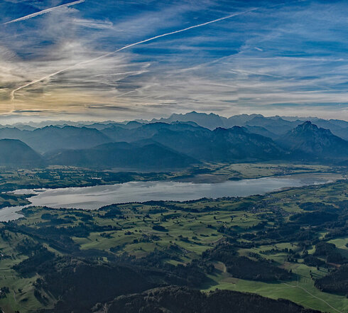 Das Foto zeigt eine Panoramaaufnahme der Alpenkette. Unzählige Gipfel reihen sich aneinander, soweit das Auge reicht. Im Vordergrund erstreckt sich die grüne Ebene mit vielen unterschiedlichen Seen und Flüssen, die eine Landschaft aus Wiesen und Wäldern durchziehen. Der Himmel ist blau, von wenigen Wolken durchzogen. Die Sonne scheint.  