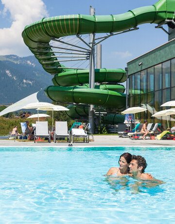 Das Bild erfasst eine lebhafte Szene in einem Resort. Der Schwerpunkt liegt auf einer großen, grünen Wasserrutsche, die gerade in Gebrauch ist. Die Rutsche ist gekrümmt und erscheint aus einem robusten Material gefertigt zu sein. Sie befindet sich in der Mitte des Bildes und zieht die Aufmerksamkeit auf sich.  Im Vordergrund schwimmen zwei Personen in einem Becken. Sie stehen einander nahe, was darauf hindeutet, dass sie vielleicht gemeinsam das Wasserrutschenvergnügen genießen. Das Becken selbst ist ein lebhaftes Blau und kontrastiert gut mit dem Grün der Rutsche.  Der Hintergrund des Bildes offenbart eine schöne Berglandschaft. Die Berge sind grün und üppig, was der Resortszenerie einen Hauch von Natur verleiht.  Das Resort selbst wird durch einen großen Gebäudekomplex mit einem grünen Dach gekennzeichnet. Dieses Gebäude ist wahrscheinlich Teil der Resorteinrichtungen und beherbergt Amenities wie ein Restaurant oder ein Geschäft.  Insgesamt zeichnet das Bild ein Bild von einem unterhaltsamen und erholsamen Tag in einem Resort, wo Menschen die Wasserrutsche genießen und die schöne Bergkulisse erfreuen.