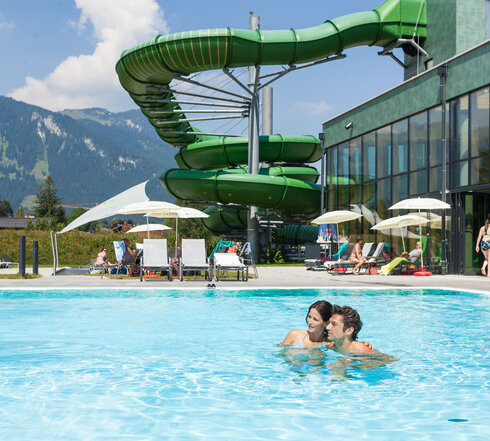 Das Bild erfasst eine lebhafte Szene in einem Resort. Der Schwerpunkt liegt auf einer großen, grünen Wasserrutsche, die gerade in Gebrauch ist. Die Rutsche ist gekrümmt und erscheint aus einem robusten Material gefertigt zu sein. Sie befindet sich in der Mitte des Bildes und zieht die Aufmerksamkeit auf sich.  Im Vordergrund schwimmen zwei Personen in einem Becken. Sie stehen einander nahe, was darauf hindeutet, dass sie vielleicht gemeinsam das Wasserrutschenvergnügen genießen. Das Becken selbst ist ein lebhaftes Blau und kontrastiert gut mit dem Grün der Rutsche.  Der Hintergrund des Bildes offenbart eine schöne Berglandschaft. Die Berge sind grün und üppig, was der Resortszenerie einen Hauch von Natur verleiht.  Das Resort selbst wird durch einen großen Gebäudekomplex mit einem grünen Dach gekennzeichnet. Dieses Gebäude ist wahrscheinlich Teil der Resorteinrichtungen und beherbergt Amenities wie ein Restaurant oder ein Geschäft.  Insgesamt zeichnet das Bild ein Bild von einem unterhaltsamen und erholsamen Tag in einem Resort, wo Menschen die Wasserrutsche genießen und die schöne Bergkulisse erfreuen.